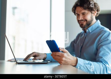 Smiling Caucasian Man Using Smartphone While Working On Laptop At Modern Office, Business Concept Stock Photo