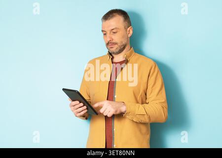Focused Caucasian Man Using Tablet Device, Standing Against Blue Background, Business Concept Stock Photo