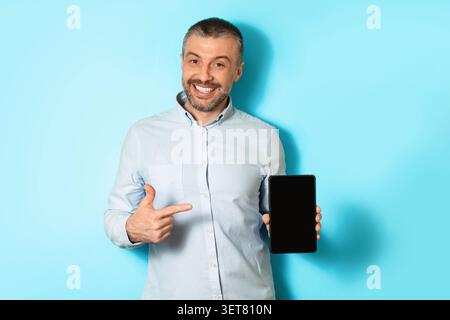 Smiling Caucasian Man Pointing At Tablet In Studio With Blue Background, Business Concept Stock Photo