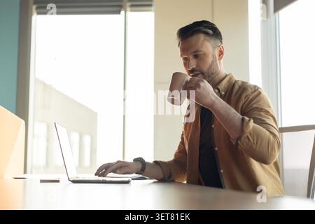 Focused Caucasian Man Working On Laptop While Drinking Coffee In Modern Office, Business Concept Stock Photo
