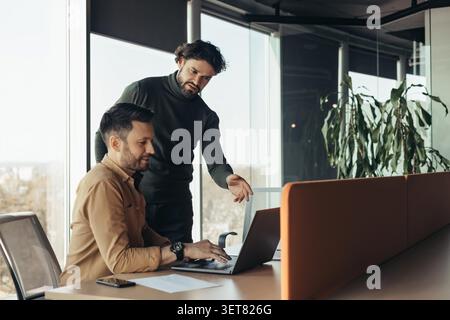 Two Men Collaborating In Office, One Explaining While Pointing At Laptop, Business Concept Stock Photo
