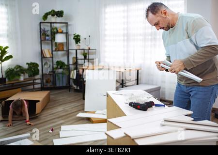 Man assembling furniture while child plays nearby in cozy home Stock Photo
