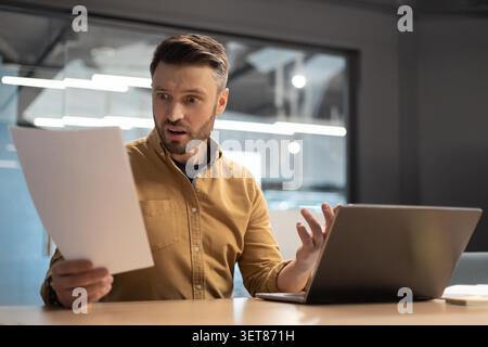Surprised Caucasian Male Office Worker Reviewing Documents With Laptop, Business Concept Stock Photo