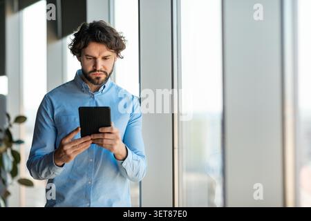Focused Caucasian Male Using Tablet While Standing By Office Window, Business Concept Stock Photo