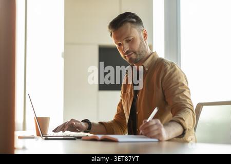 Businessman Writing Notes And Working On Laptop In Modern Office, Focused Expression Stock Photo