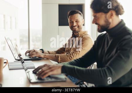 Two Smiling Men Working Together In Office, Focused On Laptops, Business Concept Stock Photo