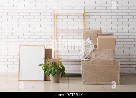 Moving day essentials: cardboard boxes, empty picture frame, houseplant, and shelf against white brick wall Stock Photo