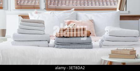 Freshly laundered towels and bedding stacks on white bed with cotton bolls and blinds in bright bedroom, the bedroom features natural light from a win Stock Photo