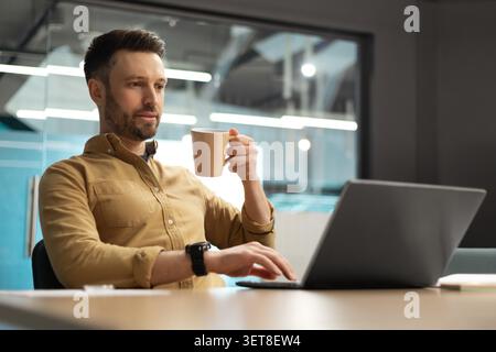 Focused White Male Working On Laptop In Modern Office, Holding Coffee Cup, Business Concept Stock Photo