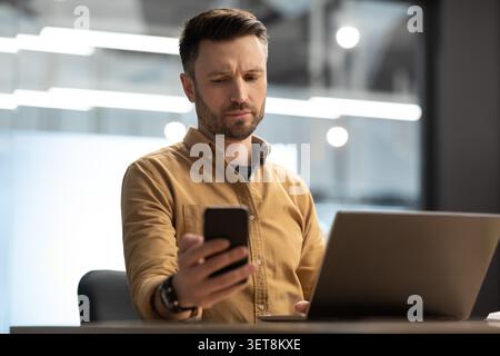 Focused White Male Professional Using Smartphone And Laptop At Office, Business Concept Stock Photo