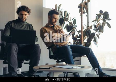 Young Caucasian Men Working Together In Office, One Using Laptop And One Using Tablet, Business Concept Stock Photo