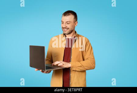 Smiling Caucasian Male With Laptop In Hand, Standing Against Blue Background, Business Concept Stock Photo