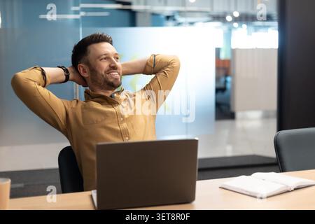 Smiling Businessman Relaxing At Desk With Laptop In Modern Office, Business Concept Stock Photo
