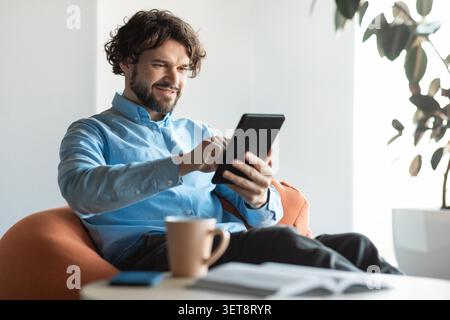 Smiling Caucasian Man Using Tablet While Relaxing In Stylish Living Room At Home Stock Photo