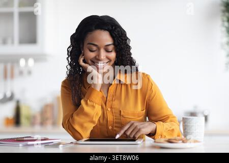Smiling Black Woman Using Tablet At Kitchen Table With Cookies And Coffee, Home Lifestyle Concept Stock Photo