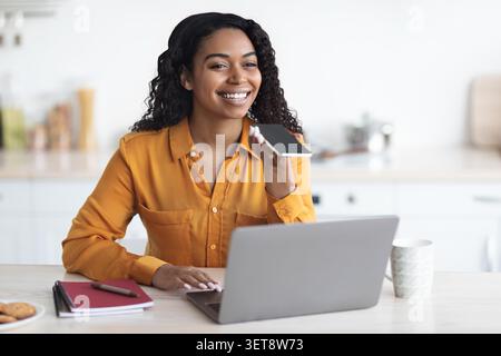 Smiling Young Black Woman Using Smartphone And Laptop At Home Office, Working Concept Stock Photo