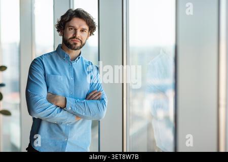Pensive Young Caucasian Man Standing By Window In Office, Business Concept Stock Photo