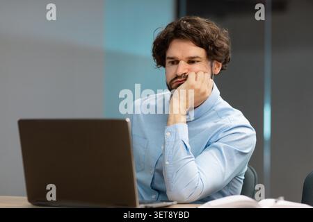 Pensive Young Caucasian Man Working On Laptop In Office, Focusing On Screen, Business Concept Stock Photo