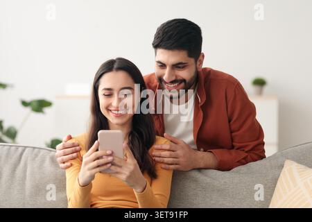 Happy Young Couple Using Smartphone Together At Home, Smiling And Enjoying Time Together Stock Photo