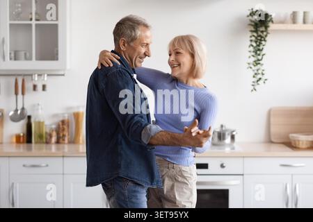 Happy Elderly Caucasian Couple Dancing Together In Kitchen, Joyful Home Lifestyle Stock Photo