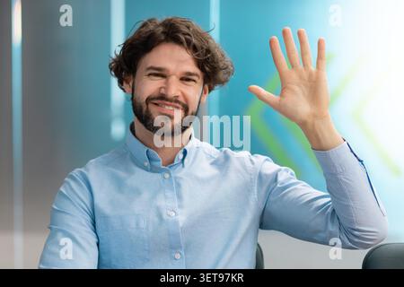 Smiling Young Man With Curly Hair Waving Hand In Office, Business Concept Stock Photo