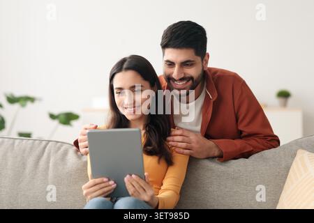 Happy Young Couple Smiling While Using Tablet At Home, Near Couch Stock Photo
