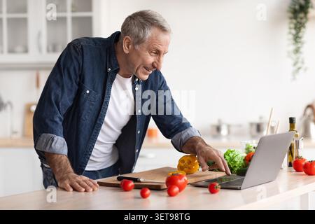Smiling Elderly Caucasian Man Cooking Healthy Meal In Kitchen, Using Laptop For Recipe Stock Photo