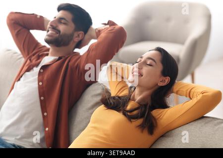 Happy Young Couple Relaxing Together On Sofa At Home, Smiling. Lifestyle Concept Stock Photo