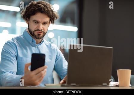 Focused Man With Curly Hair Looking At Smartphone While Working At Laptop In Office Stock Photo