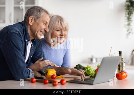 Happy Elderly Caucasian Couple Using Laptop Together In Kitchen, Cooking Concept Stock Photo