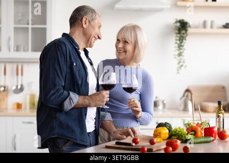 Happy Elderly Caucasian Couple Enjoying Wine In Kitchen, Preparing Healthy Meal Together Stock Photo
