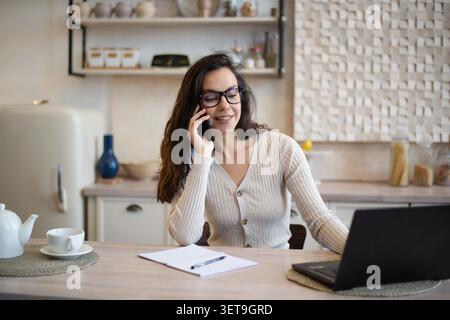 Woman Talking On Phone While Working On Laptop In Modern Kitchen, Business Concept Stock Photo