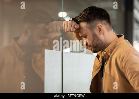 Worried Caucasian Man Leaning On Glass Wall In Office, Business Concept Stock Photo