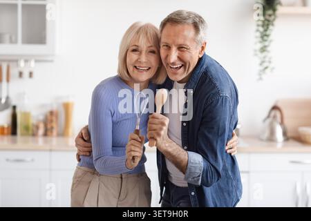 Happy Elderly Caucasian Couple Cooking Together In Kitchen, Smiling And Holding Utensils Stock Photo