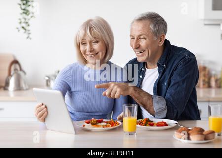Happy Senior Caucasian Couple Enjoying Meal Together And Using Tablet In Kitchen Stock Photo