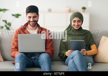 Happy Young Couple Using Laptop And Tablet Together At Home, Digital Lifestyle Concept Stock Photo