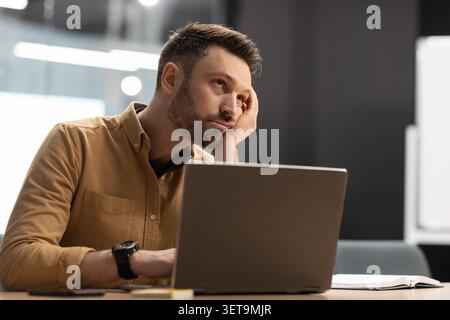Pensive Young Caucasian Man Working On Laptop At Office, Focused And Worried, Business Concept Stock Photo