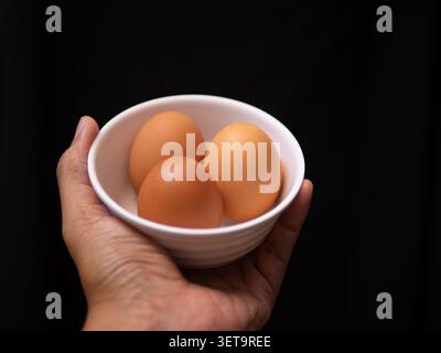 Three chicken eggs on a white bowl held by a left hand in a black background Stock Photo