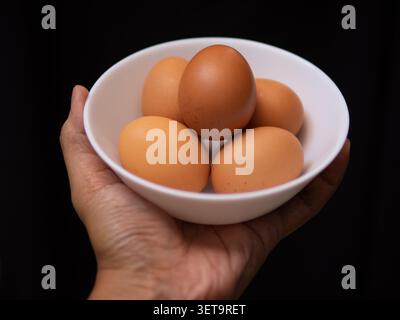 five chicken eggs on a white bowl held by a left hand in a black background Stock Photo