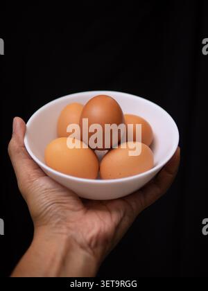 five chicken eggs on a white bowl held by a left hand in a black background Stock Photo