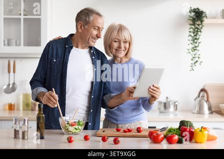 Happy Elderly Caucasian Couple Cooking Together In Kitchen, Using Tablet For Recipe, Weight Loss Concept Stock Photo