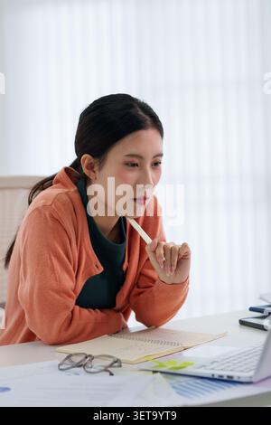 Thoughtful Asian professional woman holding pen and reviewing papers at laptop workstation in bright home office space Stock Photo