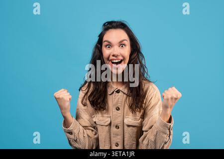 Excited Young Woman Celebrating Success With Clenched Fists, Blue Background Stock Photo