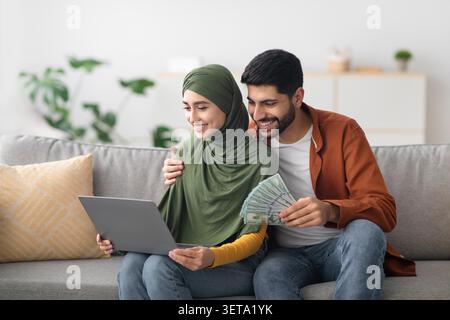 Happy Middle Eastern Couple Sitting Together At Home, Woman Using Laptop And Man Holding Money Stock Photo