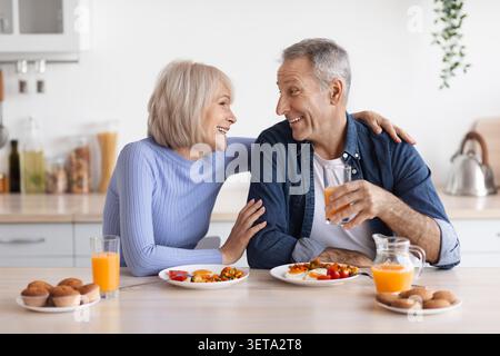 Happy Elderly Caucasian Couple Enjoying Breakfast Together In Kitchen, Smiling And Sharing Moments Stock Photo