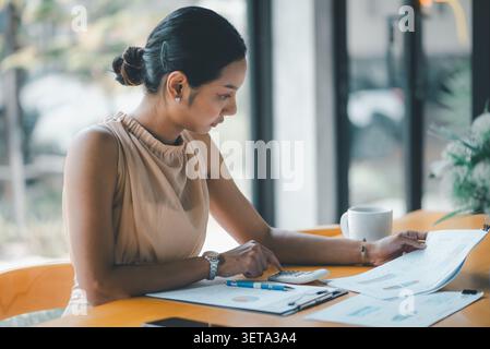 Asian businesswoman using calculator while checking financial documents, charts, and reports at a cafe desk, focused on budget planning, accounting, s Stock Photo