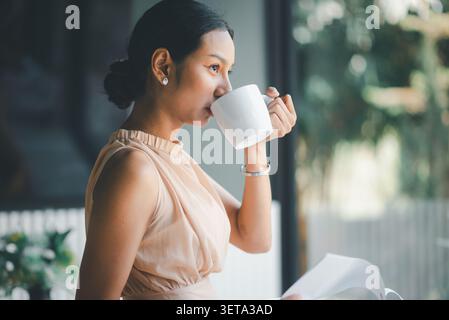 Asian businesswoman drinking coffee while holding financial papers in modern cafe office, expressing thoughtful break, budget review, accounting work, Stock Photo