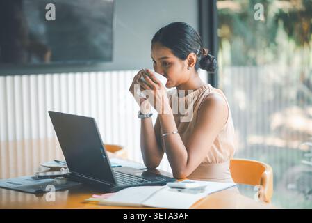 Asian businesswoman drinking coffee while working on laptop in cafe office, surrounded by reports, notebook, calculator, and earbuds, showing remote f Stock Photo