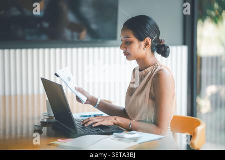 Asian businesswoman typing on laptop while holding financial report in cafe workspace, showing digital accounting, business analysis, budget planning, Stock Photo