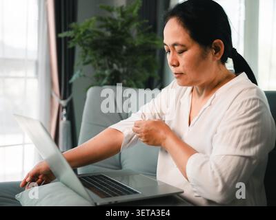 Asian woman working on a laptop at home while adjusting her sleeve, showing remote work, home office routine, online tasks, concentration, everyday co Stock Photo